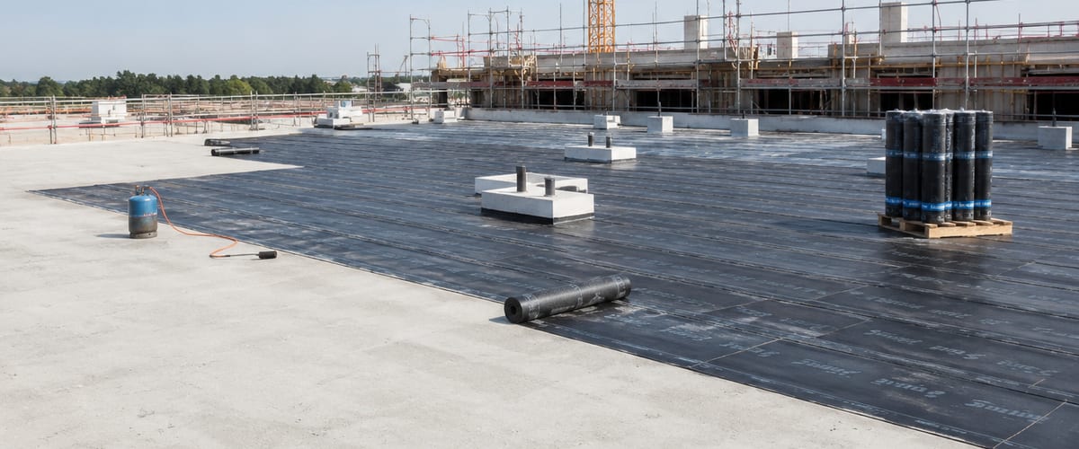 Toiture d'un bâtiment en construction : lés de bitume déroulés sur la dalle béton neuve, rouleaux en attente, grue et échafaudages en arrière-plan